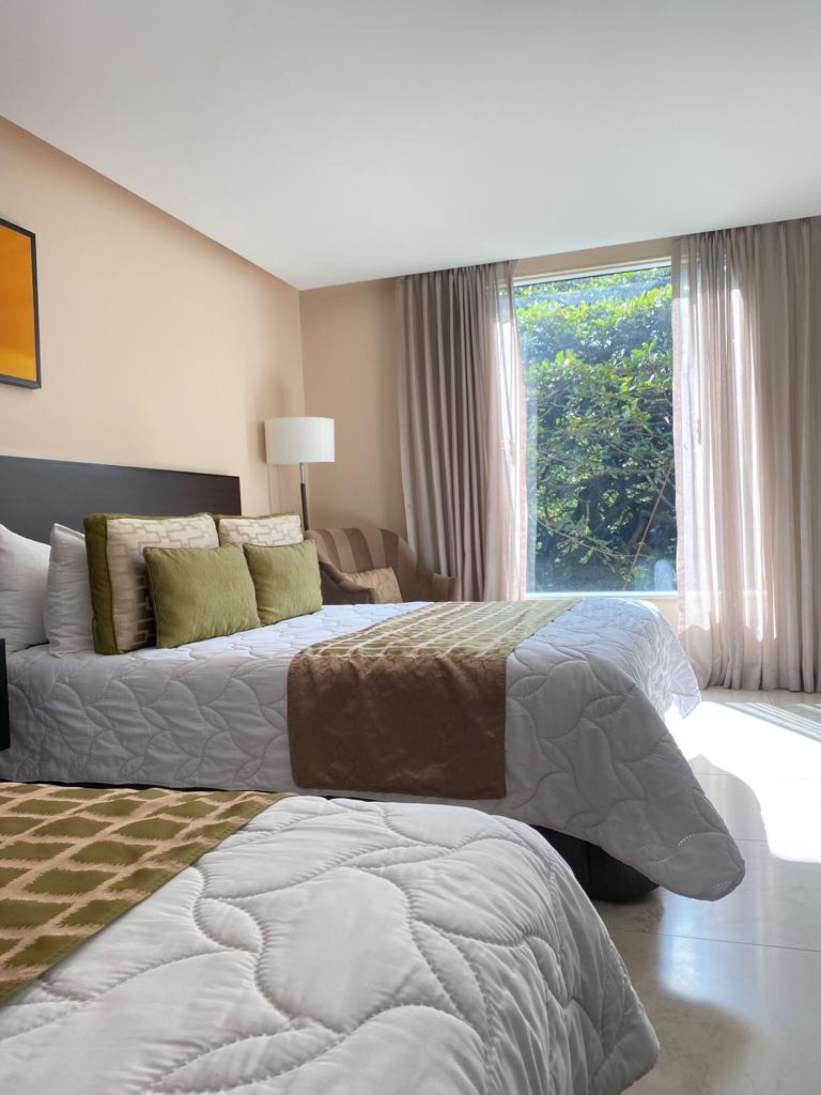 Een hotelkamer met een groot bed, een nachtkastje met een lamp, en uitzicht op groen buiten het raam.