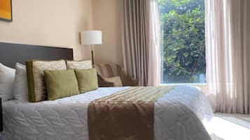 Een hotelkamer met een groot bed, een nachtkastje met een lamp, en uitzicht op groen buiten het raam.