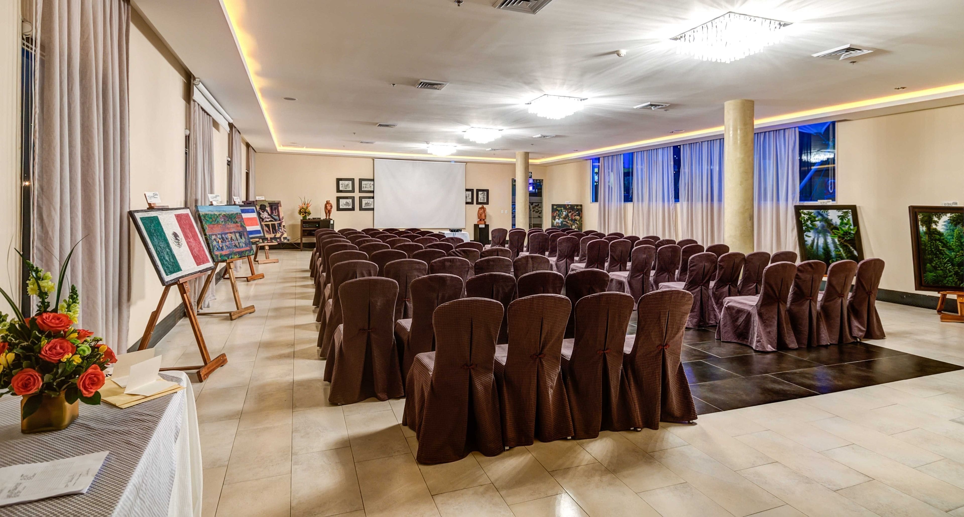 Un salón de conferencias con filas de sillas, un escenario con un caballete y cuadros en las paredes.