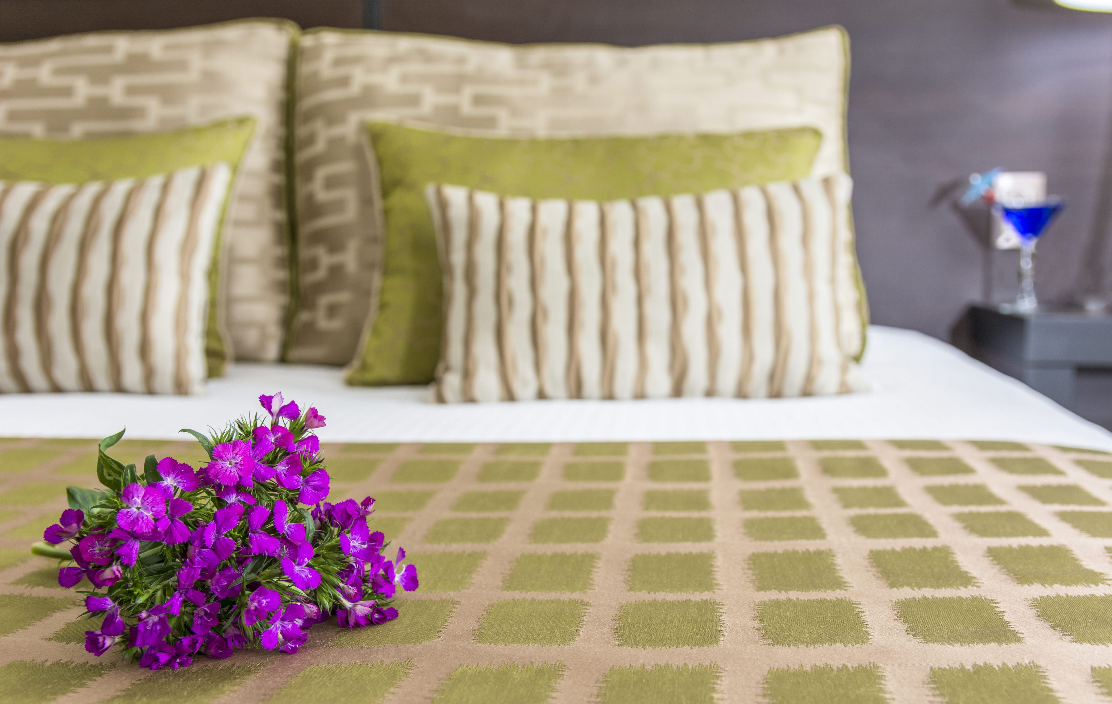 Een net opgemaakt bed met een bedsprei met een patroon, twee kussens met een geometrisch ontwerp, en een bos paarse bloemen op het bed.