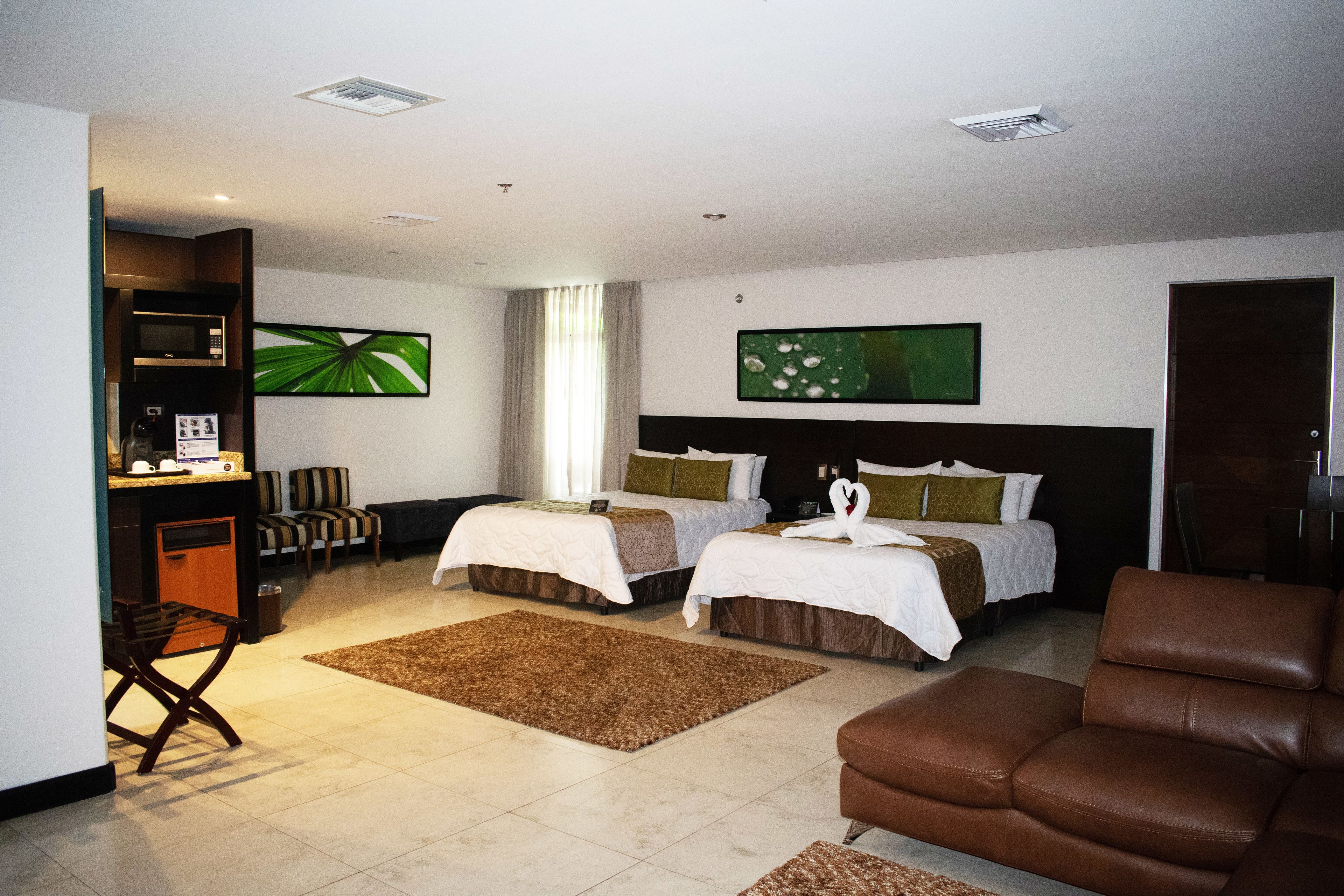 A hotel room with two beds, a brown leather sofa, a small kitchen area with a microwave, and a large window with curtains.