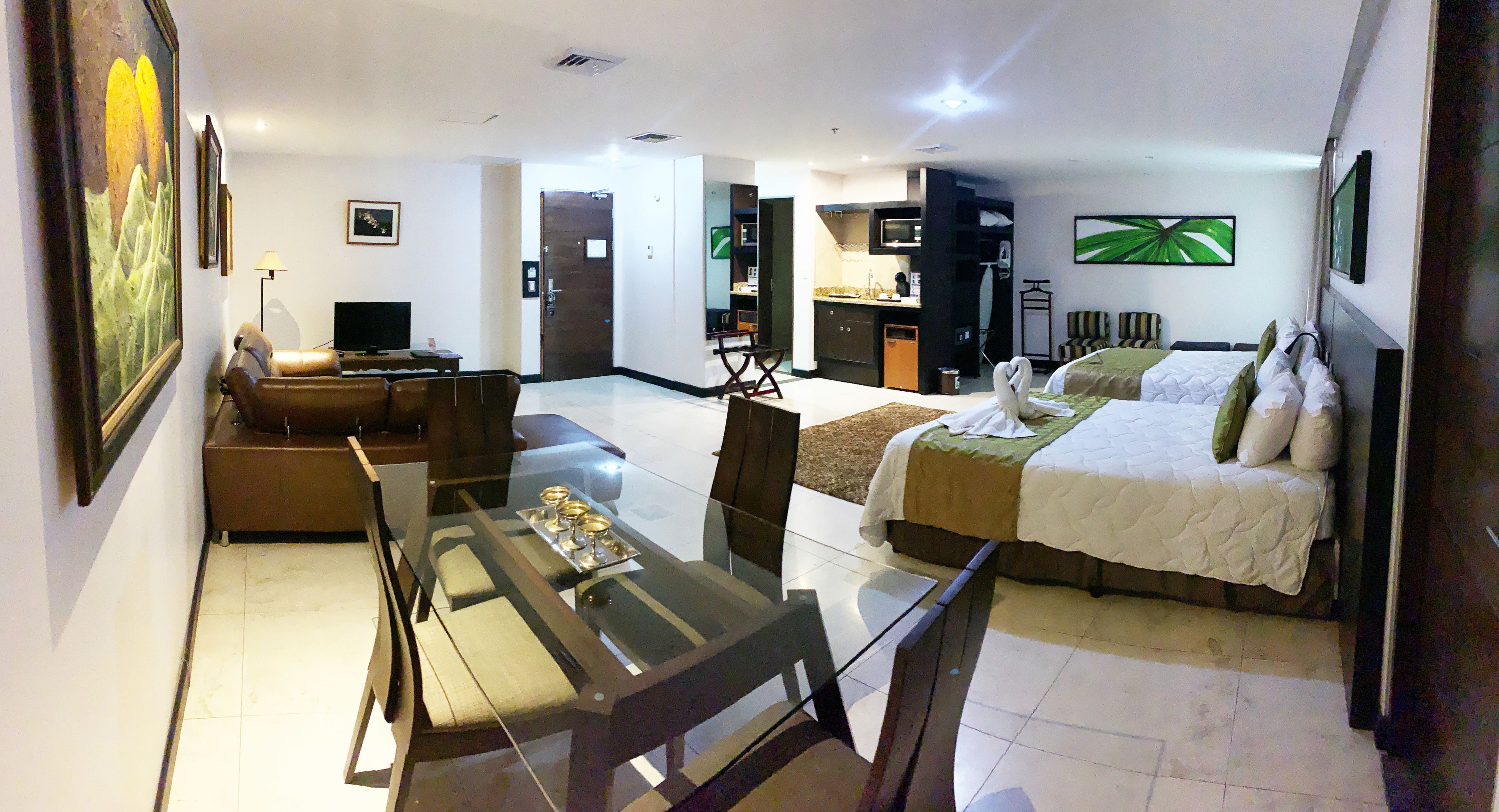 A hotel room with a bed, a sofa, a glass dining table, and a kitchen area.