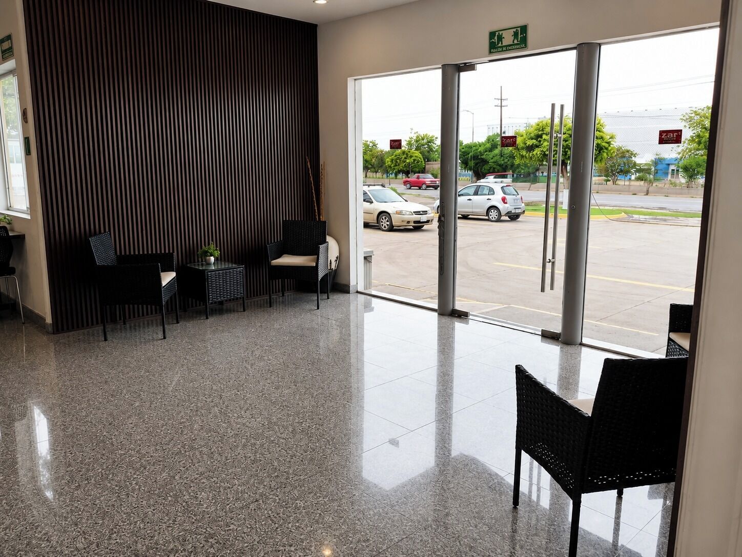 Una moderna zona de recepción con un puerta de cristal que da al exterior, un área de descanso con sillas y una mesa, y un panel de madera vertical en la pared.