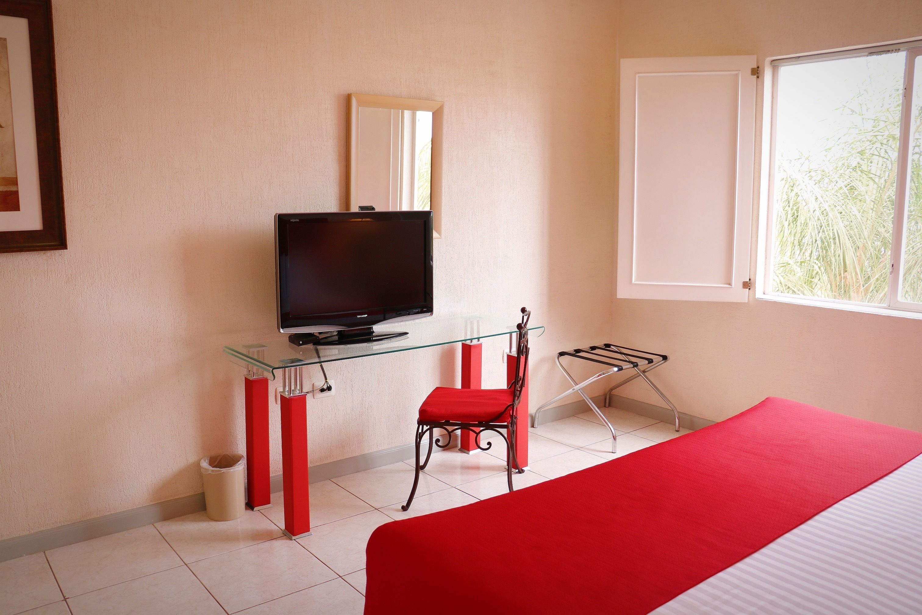 A hotel room with a bed, a television on a glass table, a red chair, a window, and a door.