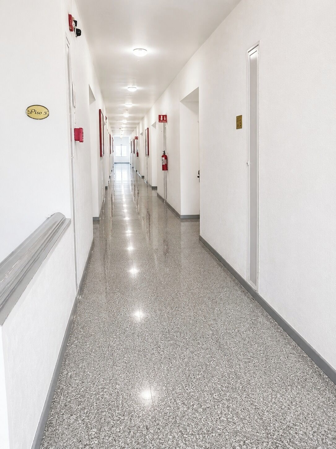 A long, narrow hallway with white walls, doors on both sides, and a polished floor.