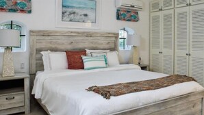 A bedroom with a wooden headboard, white bedding, and a patterned throw blanket. There are two bedside tables with lamps, a window, and a set of white closets. - Glitter Bay (Holetown)
