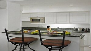 A modern kitchen with white cabinets, a microwave, and a granite countertop. There are two bar stools with cushioned seats. - Glitter Bay (Holetown)