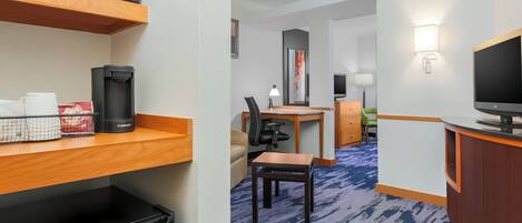 A hotel room with a kitchenette, a desk, a chair, a television, and a blue carpeted floor.