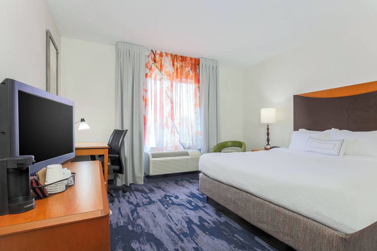 Een hotelkamer met een bed, een bureau met een computer, een televisie, een lamp en een raam met gordijnen.
