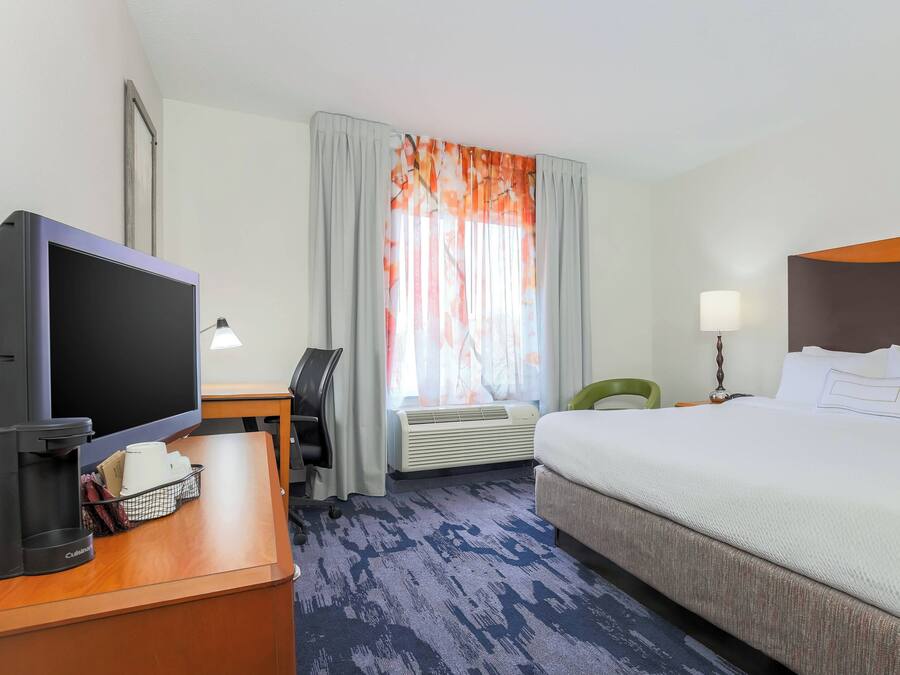 A hotel room with a bed, a desk with a computer, a TV, a lamp, and a window with curtains.