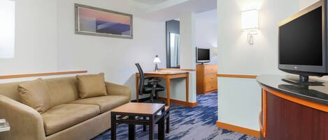 A hotel room with a beige sofa, a wooden desk with a chair, a television, and a framed picture on the wall.