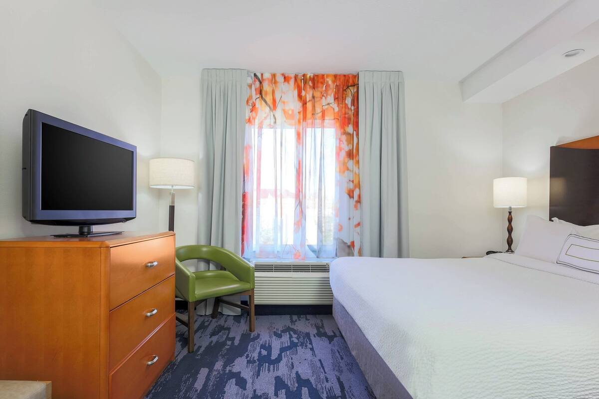 Hotelkamer met een bed, een televisie op een houten dressoir, een groene stoel en een raam met gordijnen.