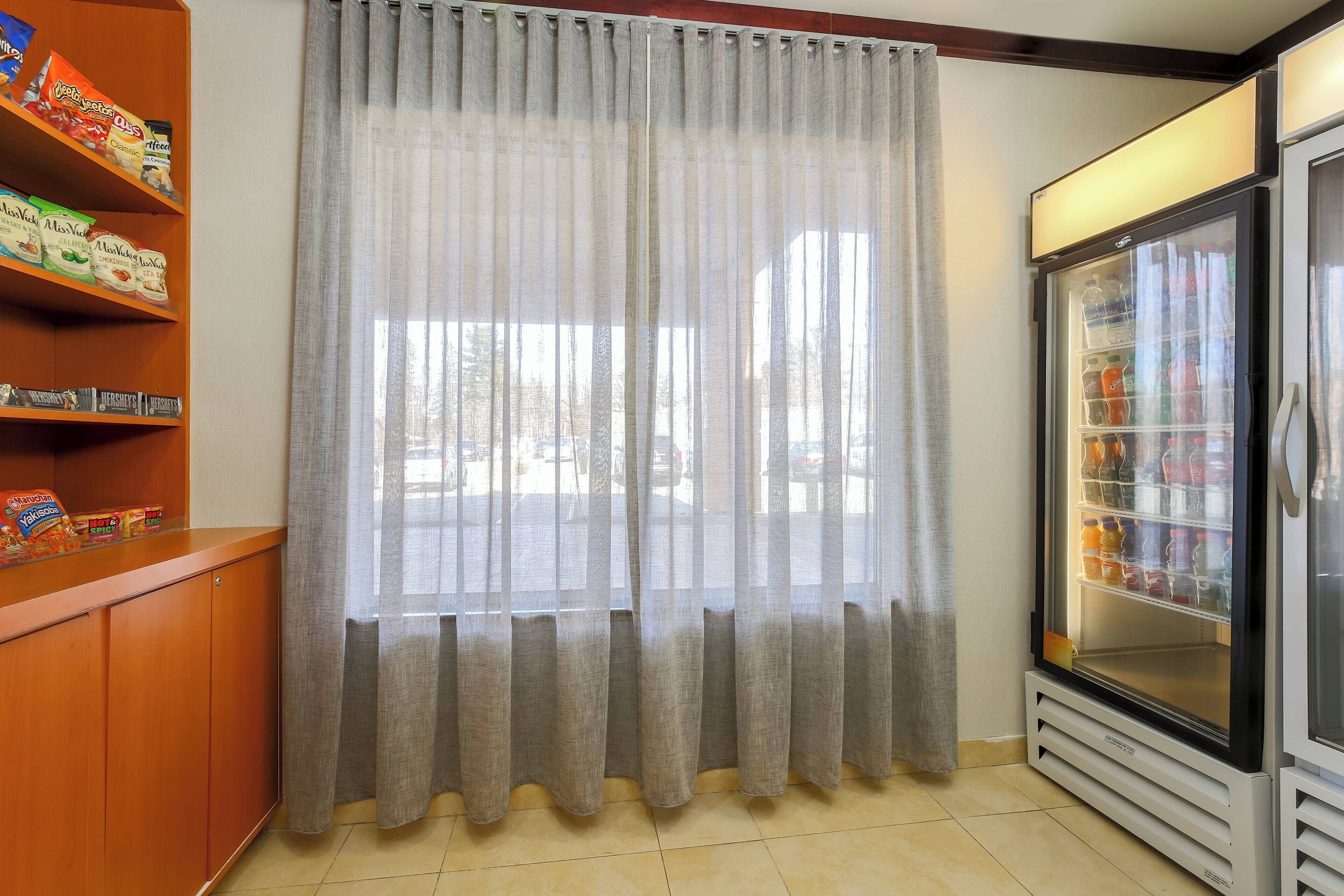 A room with a window covered by sheer curtains, a refrigerator stocked with beverages, and shelves with snacks and chocolates.