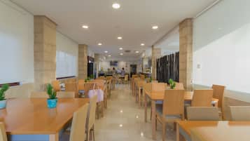 A spacious, well-lit dining area with multiple wooden tables and chairs, potted plants, and a modern interior design.