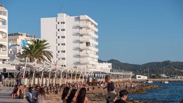 Un paseo marítimo en la playa con gente sentada y caminando, un edificio blanco con varios balcones, y una vista despejada del mar y las colinas al fondo.