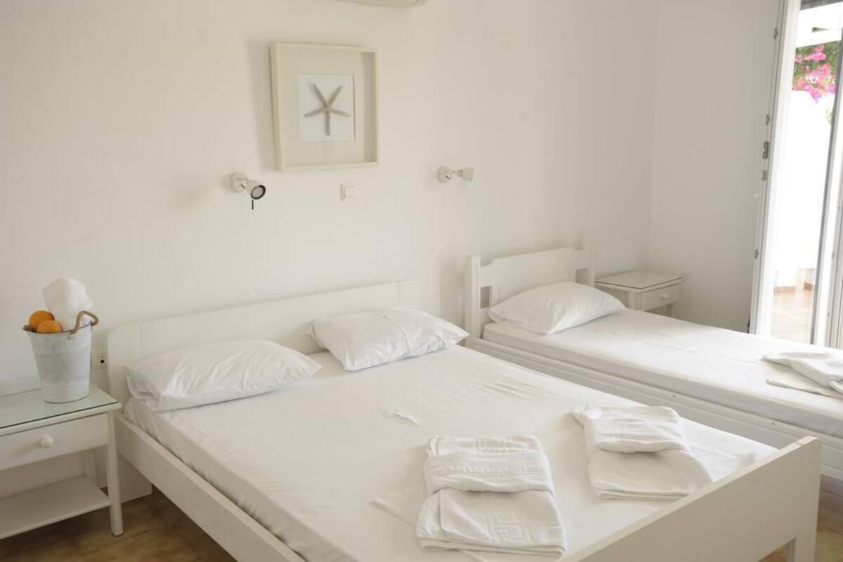Une chambre propre et blanche, avec deux lits simples, une petite table de chevet avec une lampe et un tableau encadré au mur.