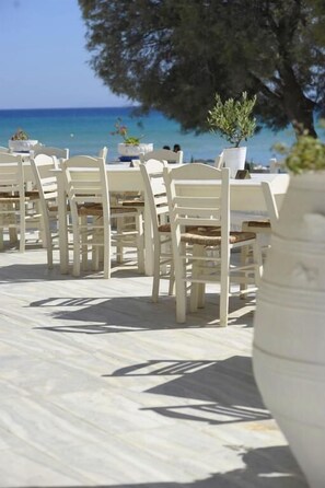 An outdoor dining area with white chairs and tables, potted plants, and a view of the sea.