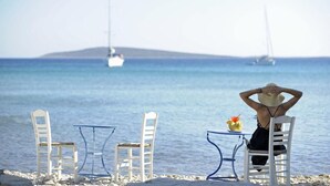 A beach scene with a person sitting at a table, two white chairs, and a table with a drink.