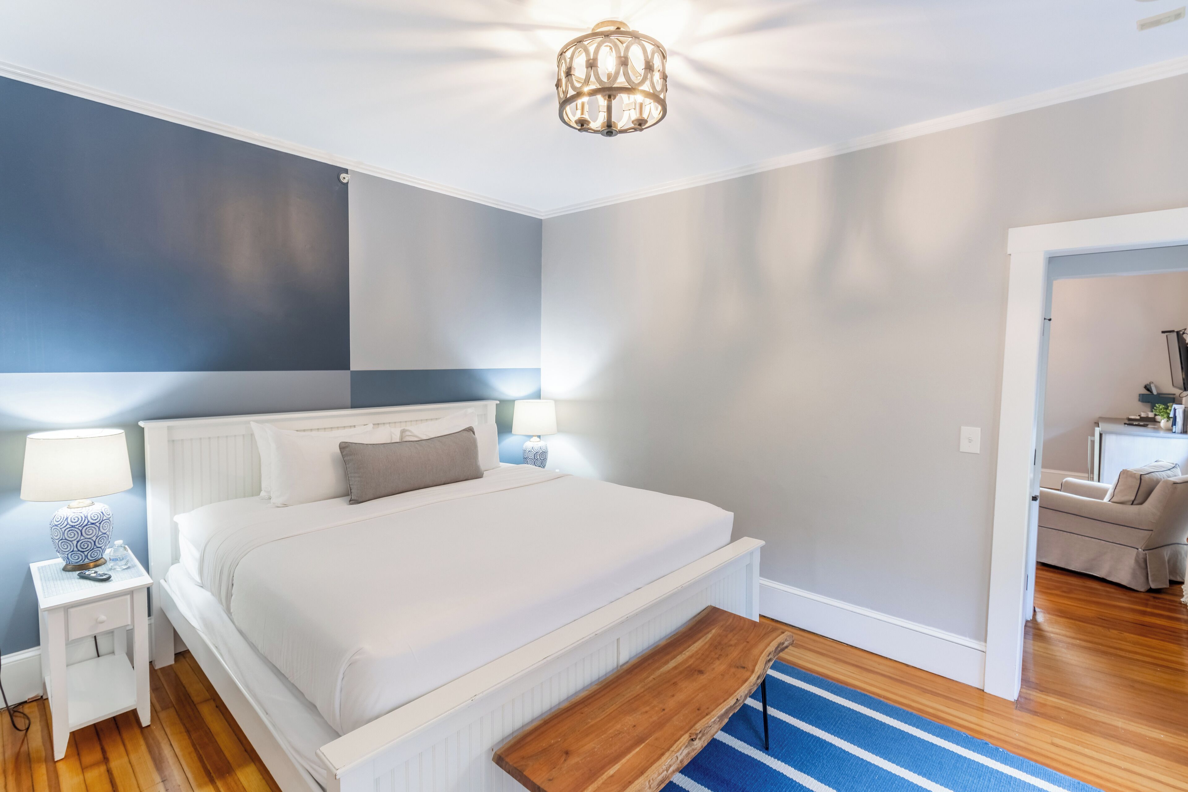 Ein Schlafzimmer mit einem weißen Bett, Nachttischen mit Lampen, einer Holzbank und einem Teppich mit blauen Streifen.