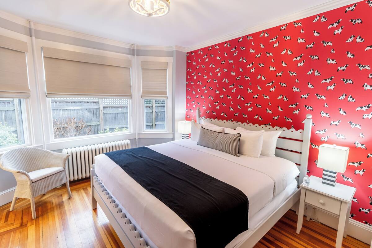 A bedroom with a large bed, a white chair, a radiator, and a window with blinds.