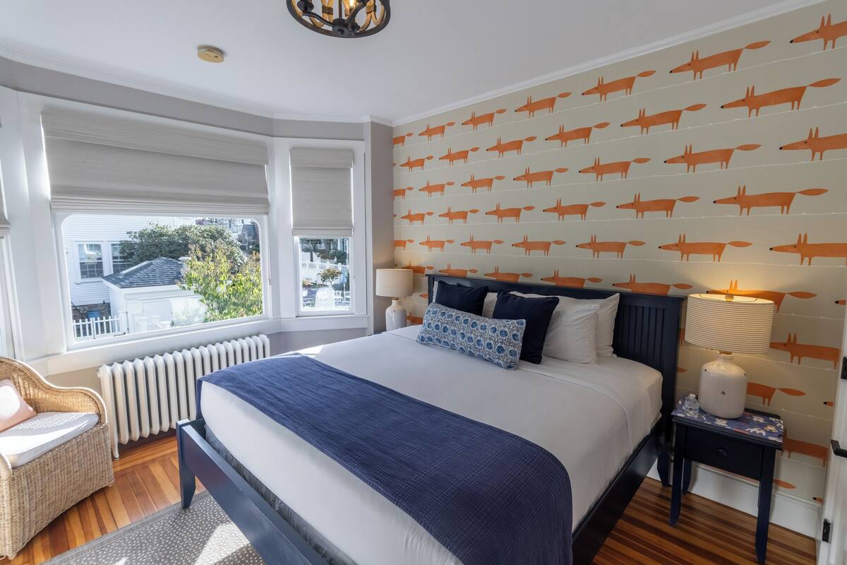 A neatly arranged bedroom with a large bed, a window with blinds, a wicker chair, and a wall with a fox pattern.