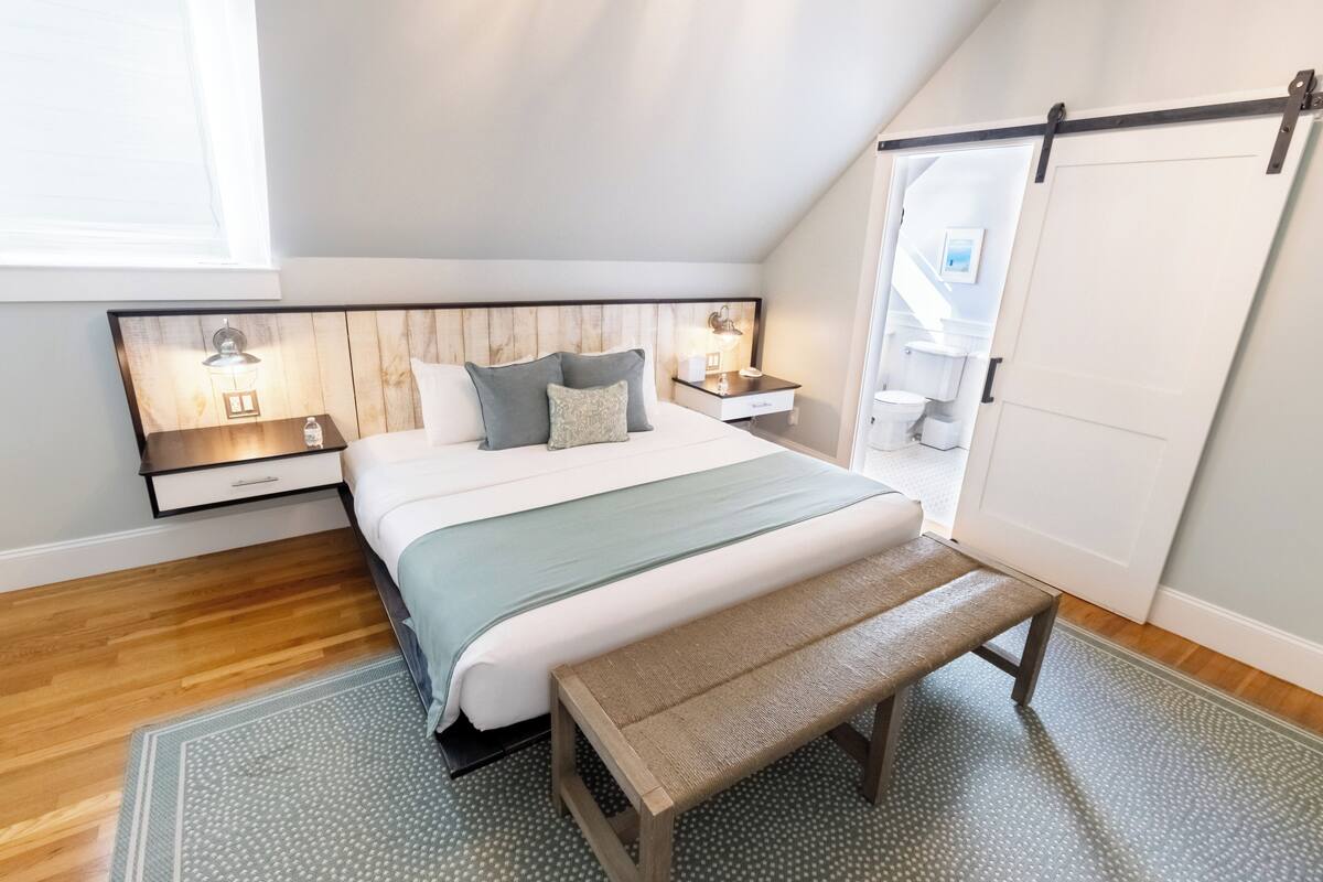 A bedroom with a wooden headboard, a large bed with a green and white bedspread, two bedside tables with lamps, a bench, and a sliding door.