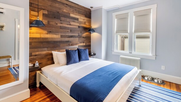 A bedroom with a wooden headboard, a bed with white and blue bedding, a blue and white striped rug, a window with blinds, and a radiator. - Paws on Pelham (Newport)