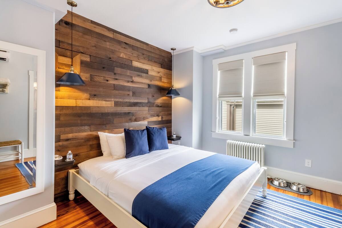 A bedroom with a wooden headboard, a bed with white and blue bedding, a blue and white striped rug, a window with blinds, and a radiator.