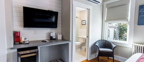 A compact hotel room with a flat-screen TV, kitchenette, wooden flooring, a window with blinds, and a view of the outside.