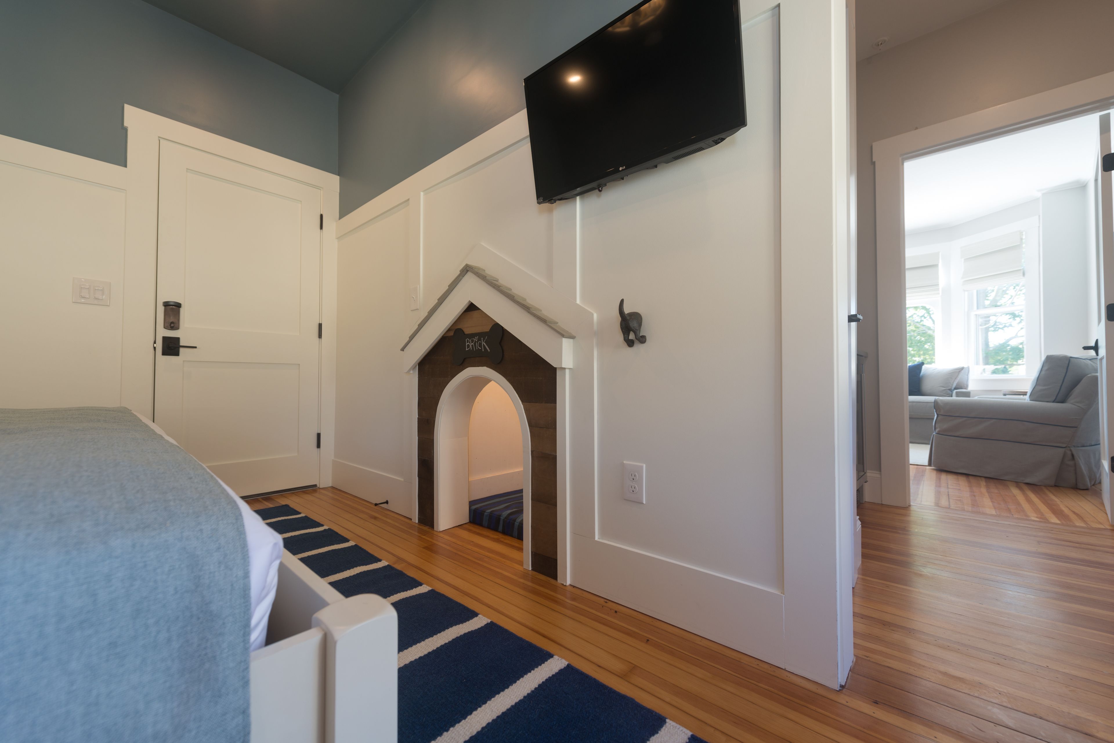 Un dormitorio con cama, televisión montada en la pared, piso de madera y una puerta que da a otra habitación.