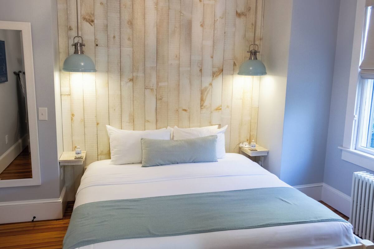 A bedroom with a wooden headboard, a bed with white linens, two bedside lamps, and a window with blinds.