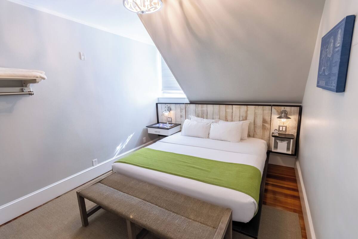 A modern bedroom with a large bed, a bench, a nightstand with a lamp, and a wall-mounted shelf.