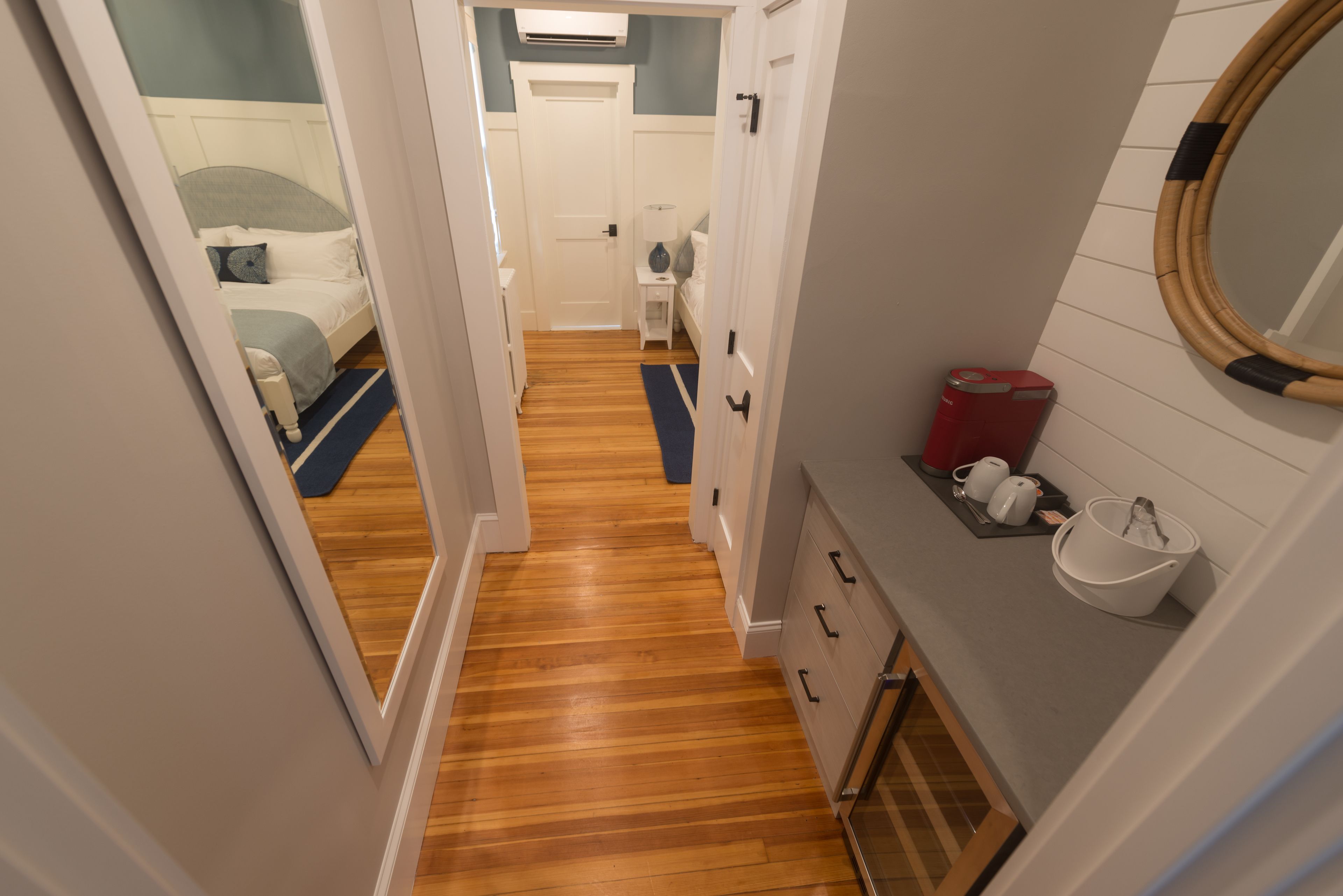 Un couloir étroit, doté d’un plancher en bois, mène à une chambre à coucher où se trouvent un lit et une table de chevet. Une petite salle de bain avec un lavabo et un miroir est visible sur la droite.