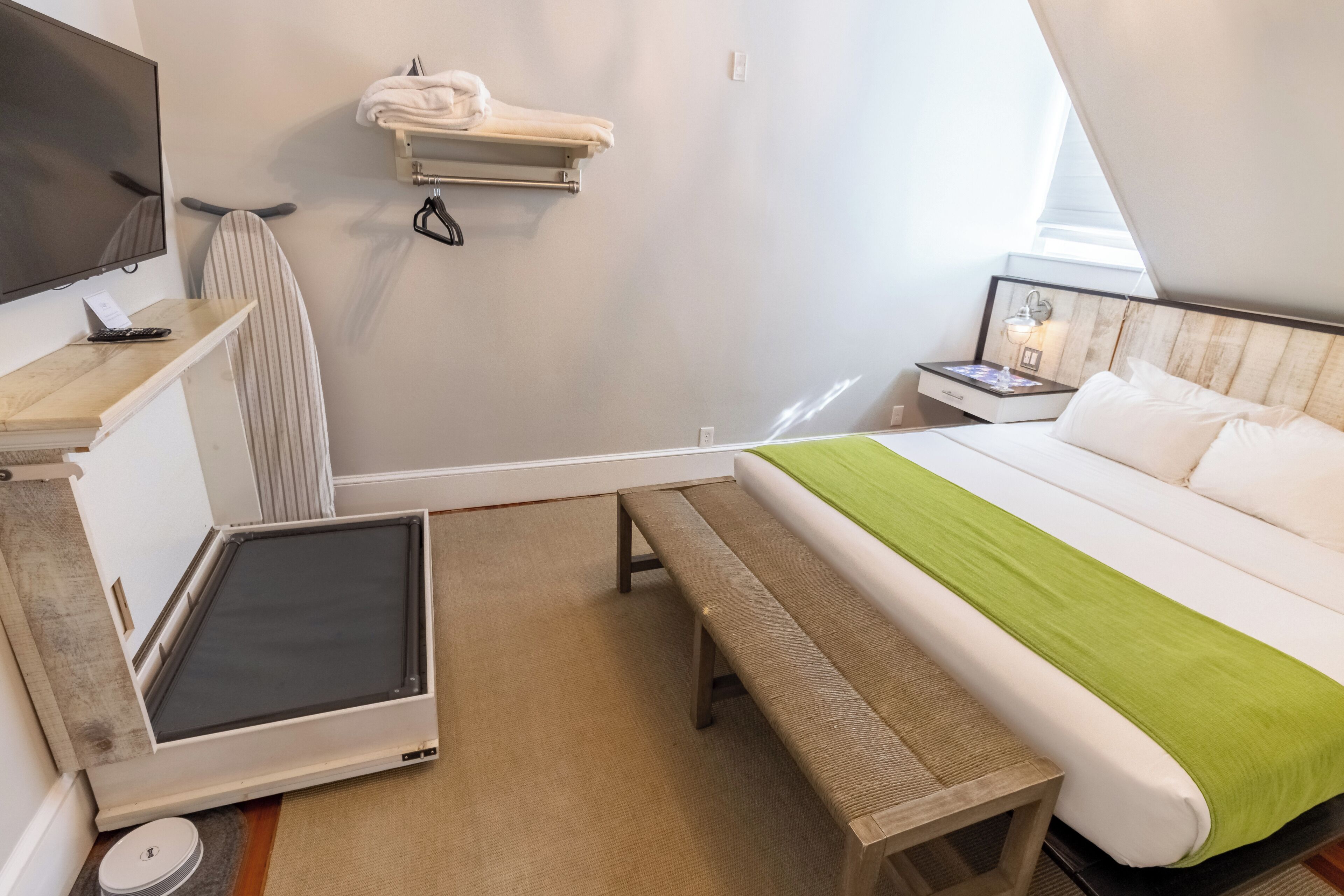 Habitació d'hotel amb llit, una panxa, televisió, prestatges per tovalloles i una taula petita amb comandament a distància.