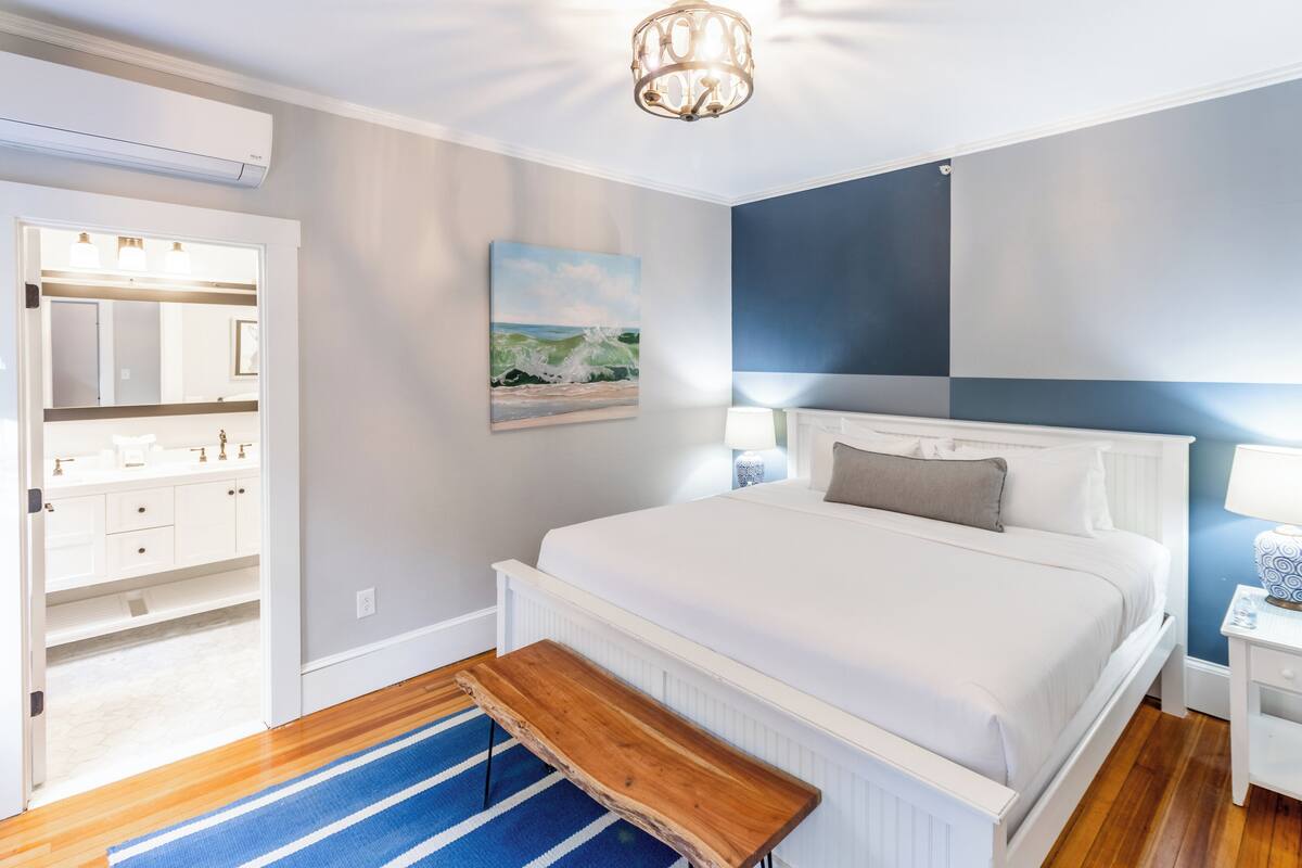 A bedroom with a large bed, a wooden bench, a nightstand, a lamp, and a painting on the wall.