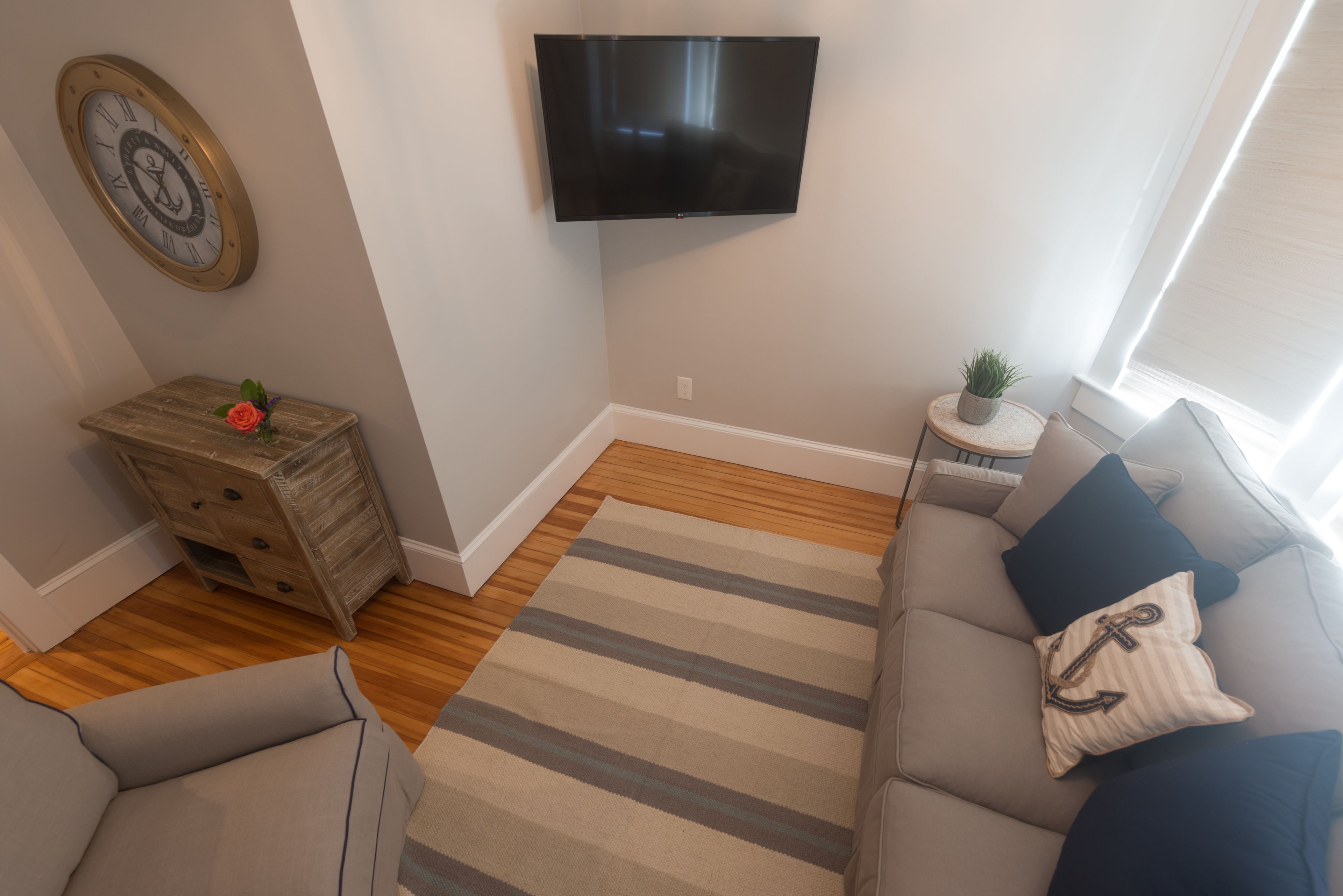 A living room with a flat-screen TV mounted on the wall, a wooden chest of drawers, a grey sofa with a striped rug, and a small side table with a plant.