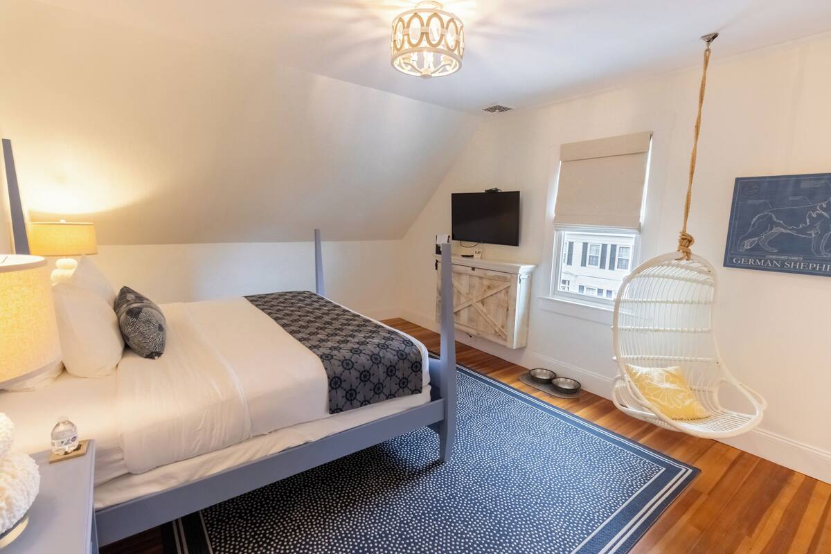 A bedroom with a bed, a hanging chair, a television, and a window with a view of buildings.