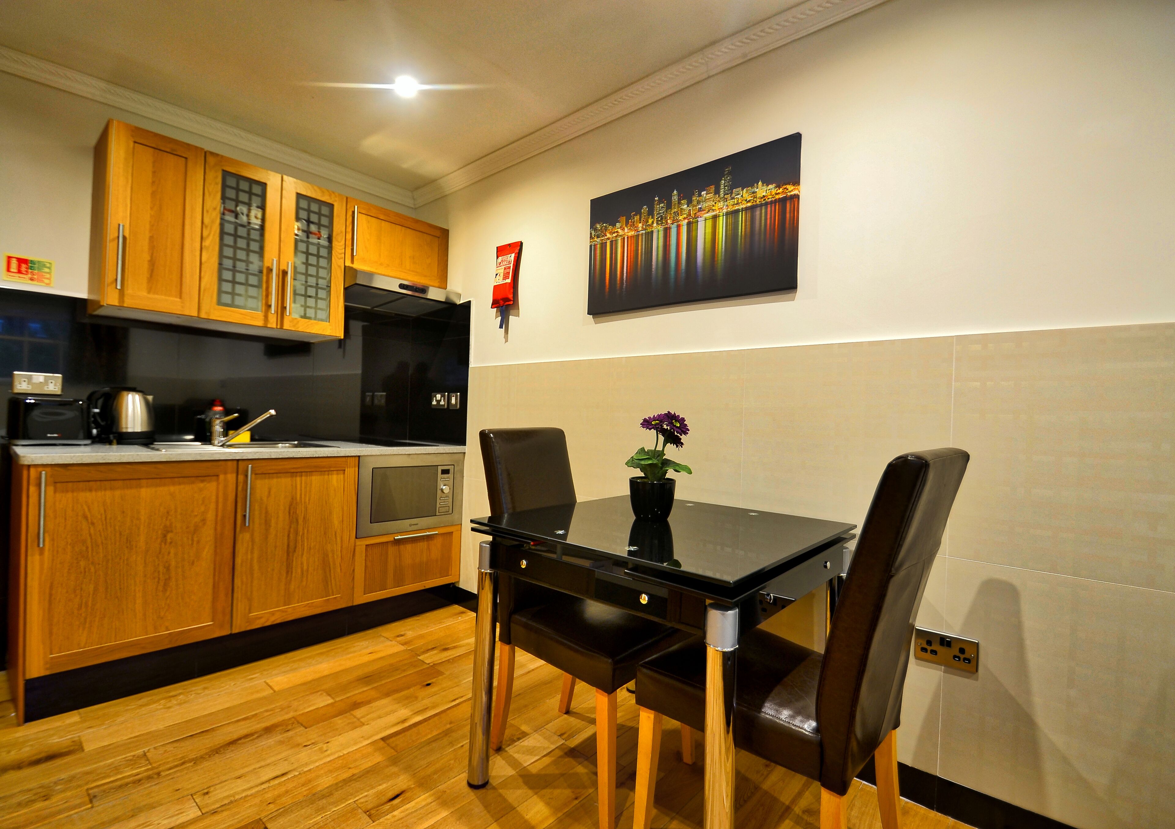 A modern kitchen with wooden cabinets, a black dining table, and a cityscape painting on the wall.