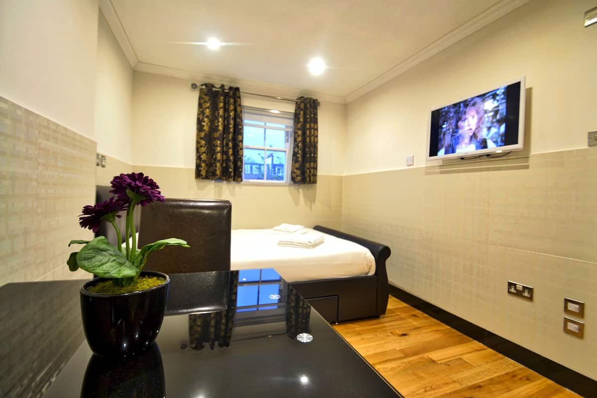 A hotel room with a bed, a TV mounted on the wall, a window with curtains, and a small table with a flower vase.