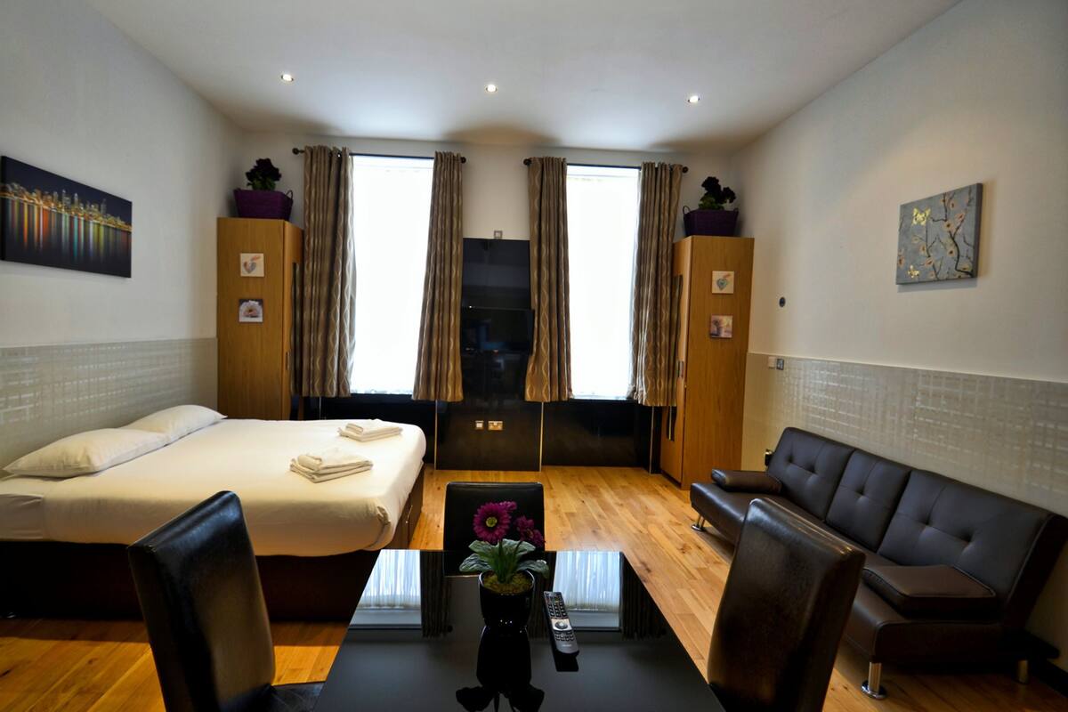A hotel room with a bed, a sofa, a dining table with chairs, a TV, and a window with curtains.