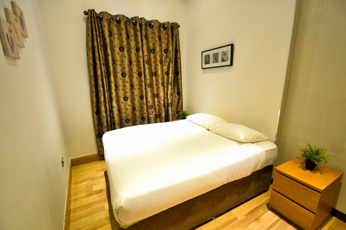 A neatly made bed with a white comforter and pillows, a wooden nightstand with a plant, and a curtain with a floral pattern.