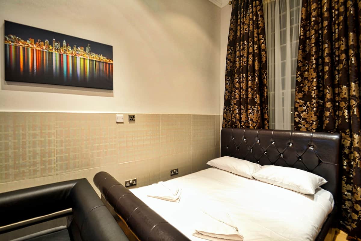 A hotel room with a bed, a large window with curtains, a cityscape painting on the wall, and a tiled wall.