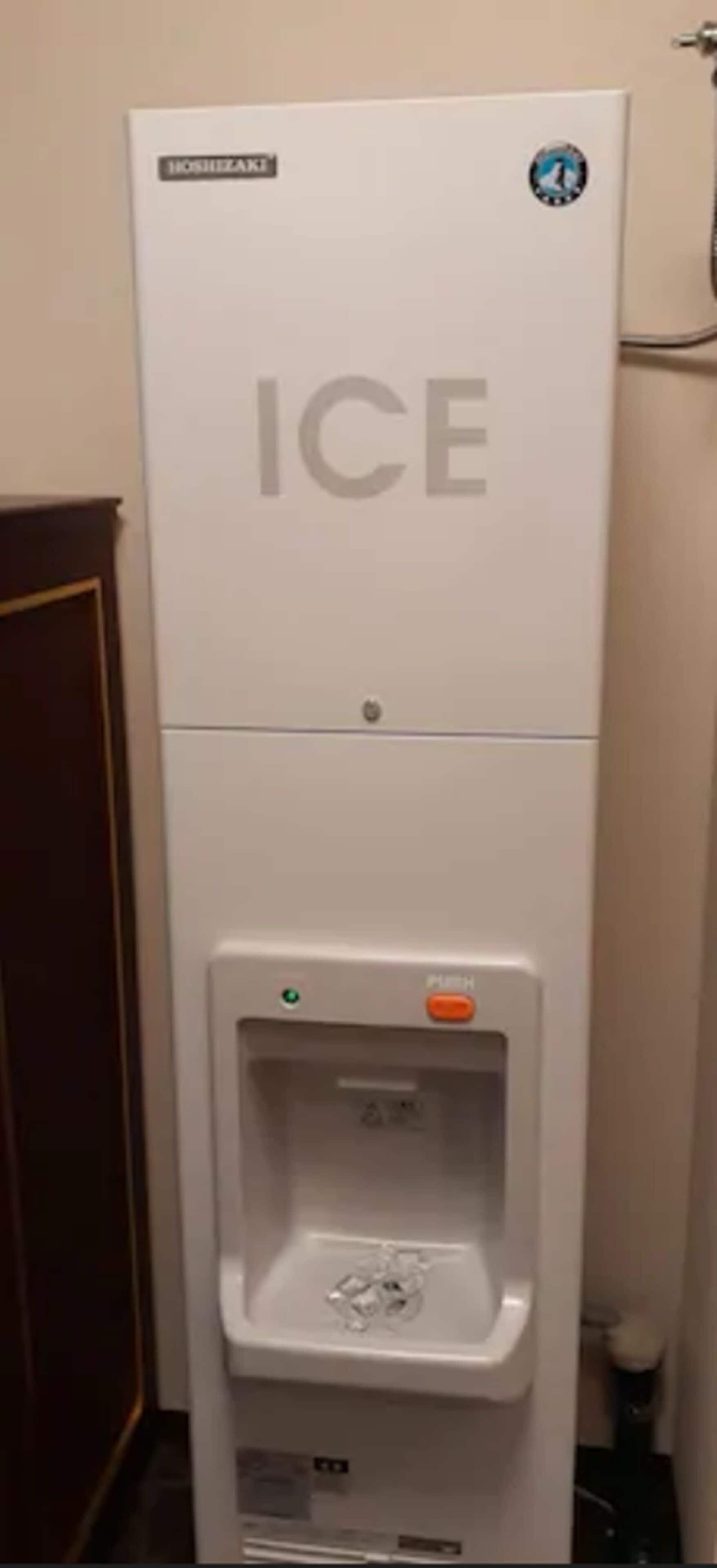 Habitación de hotel con un minibar blanco, un dispensador de agua y una puerta a la izquierda.