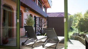 Puerta de cristal que da a un área de estar al aire libre con dos sillas y una mesa, una cerca decorativa y un edificio con tejado inclinado al fondo.