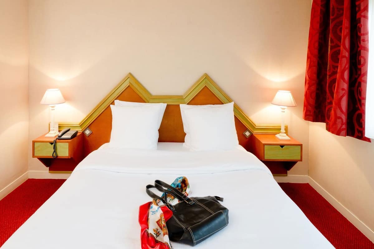 A hotel room with a bed, two bedside tables, lamps, a red curtain, and a bag on the bed.