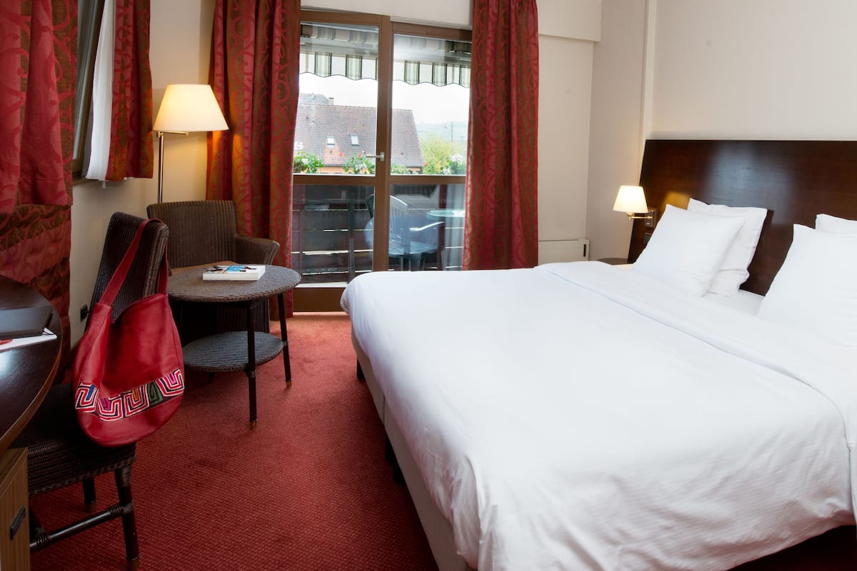 A hotel room with a large bed, two bedside lamps, a desk with a chair, a red bag, and a view of the outside through a window with curtains.