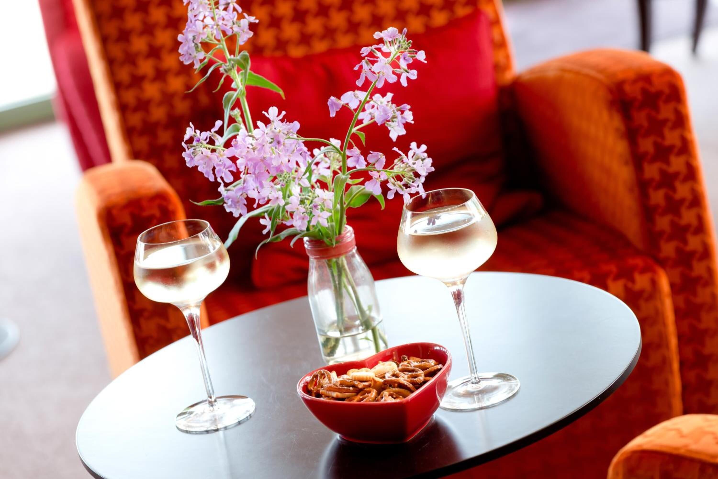 Una mesa con dos copas de vino blanco, un bol de aperitivos y un florero con flores.
