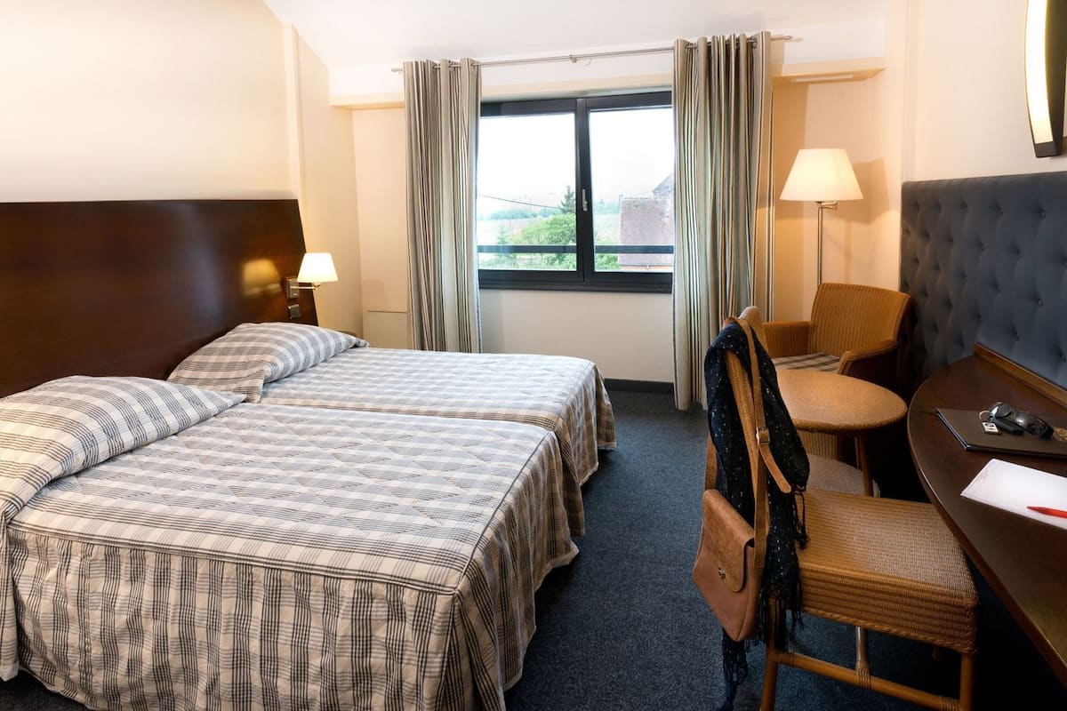 A hotel room with two beds, a desk, a chair, a lamp, and a window with curtains.