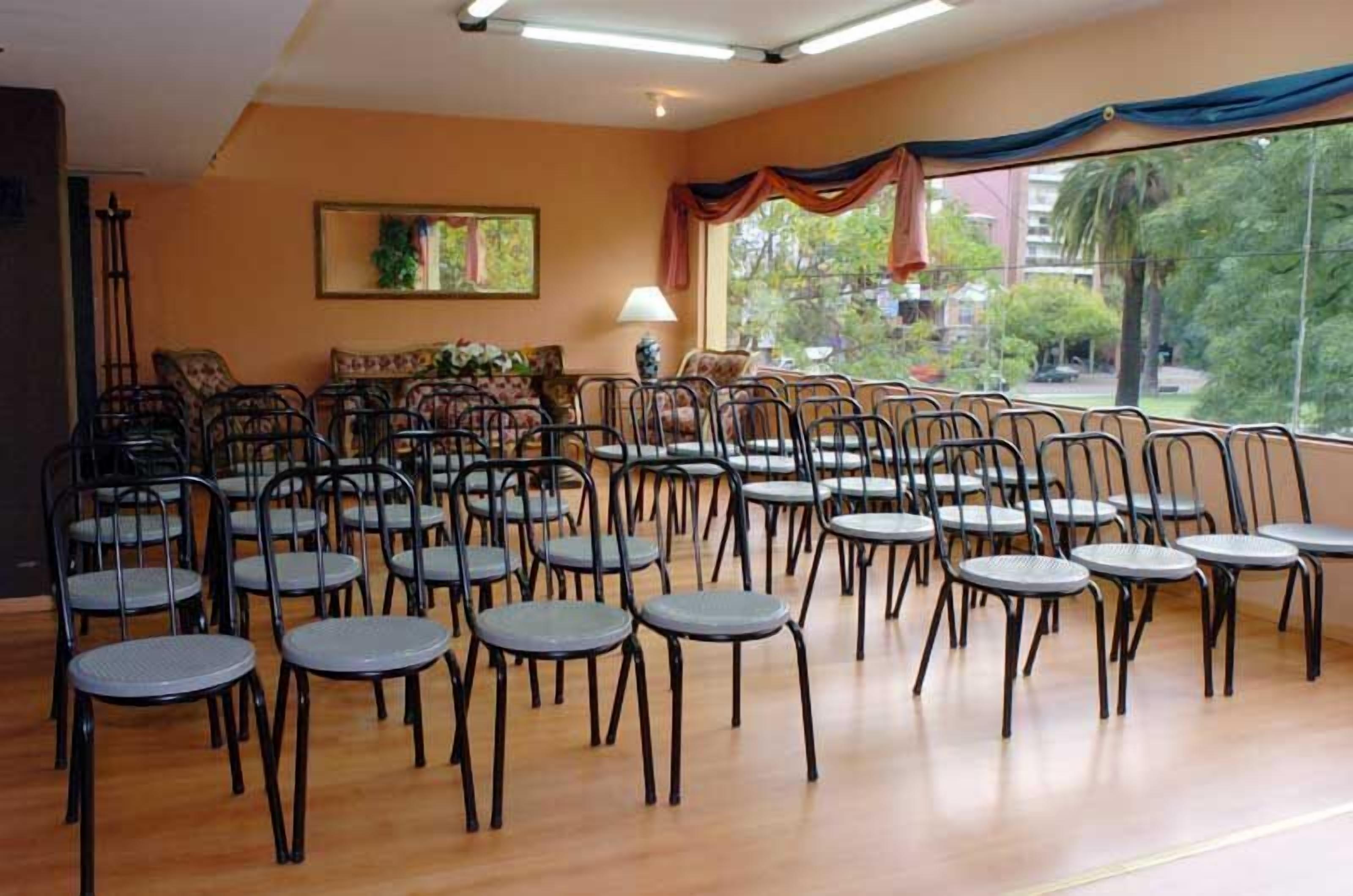 Um quarto com fileiras de cadeiras e mesas, janelas amplas com cortinas e um quadro na parede.