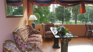A room with a sofa, armchairs, a table with a vase, and a view of trees and buildings through large windows. - Hotel del Rey (La Plata)